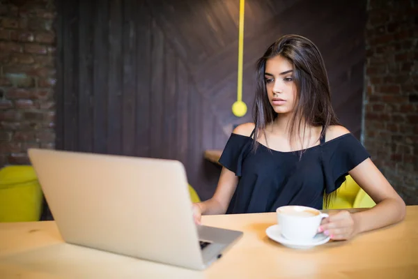 Young woman sitting in coffee shop at wooden table, drinking coffee and using smartphone. On table is laptop. Girl browsing internet, chatting, blogging. — Stock Photo, Image