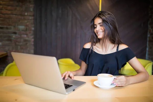 Young woman sitting in coffee shop at wooden table, drinking coffee and using smartphone. On table is laptop. Girl browsing internet, chatting, blogging. — Stock Photo, Image