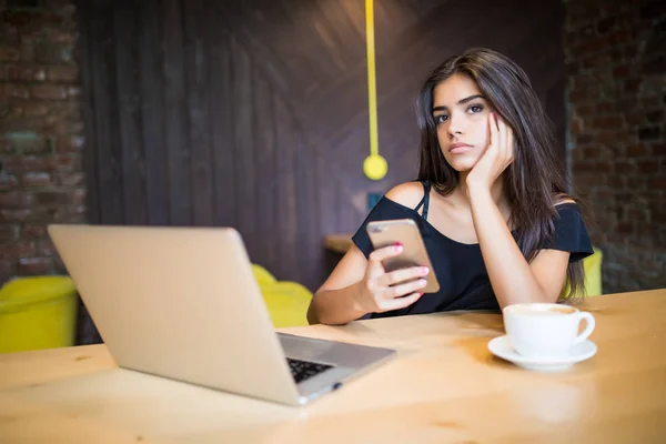 Atractiva freelancer femenina sostiene el teléfono inteligente mientras se sienta en la mesa de madera frente a la computadora abierta en la cafetería moderna. mujer creativa joven trabaja en el ordenador portátil mientras desayuna — Foto de Stock