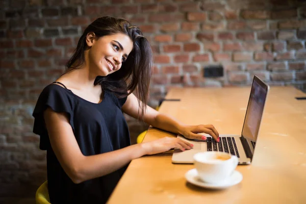 Mujer joven e independiente sentada en la cafetería con laptop y tomando café. Mujer joven con portátil en la cafetería . — Foto de Stock