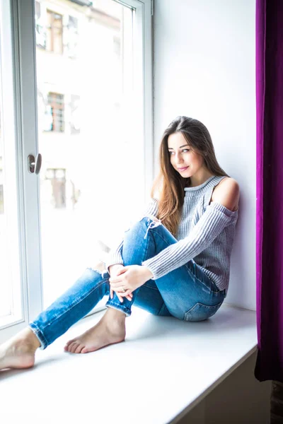 Gelukkig jongedame zittend op een vensterbank thuis en op zoek naar buiten — Stockfoto