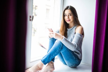 Cep telefonu evde pencere pervazına üzerinde oturan güzel genç kadın