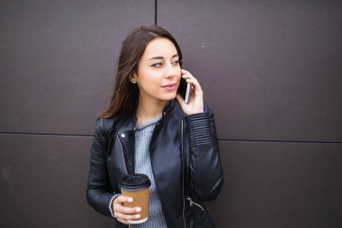 Akıllı telefonda konuşurken ve paket servisi olan restoran kahve papercup, onu muhatap ilgi ile dinlerken tutan duvara yaslanmış güzel öğrenci kız gülümseyerek