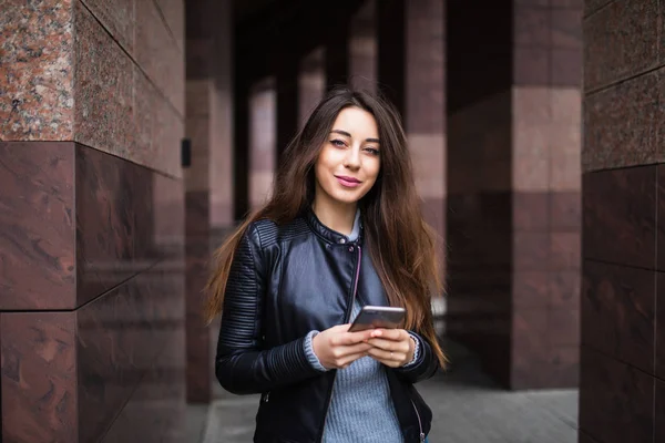 Hermosa joven está utilizando una aplicación en su dispositivo de teléfono inteligente para enviar un mensaje de texto delante de un fondo del edificio — Foto de Stock
