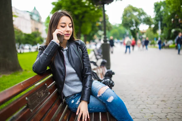 Retrato de una mujer feliz sentada en el banco y hablando por teléfono al aire libre — Foto de Stock