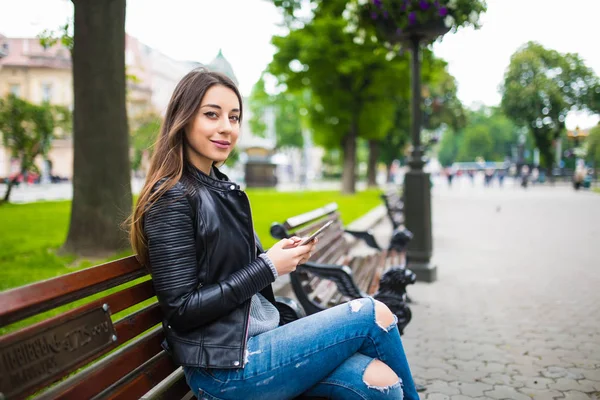 Hermosa joven sentada en el banco al aire libre y escribiendo en el teléfono — Foto de Stock