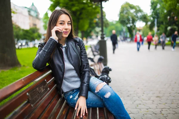 Joven hermosa mujer sentada en un banco en el parque de la ciudad y hablando por teléfono y sonriendo — Foto de Stock