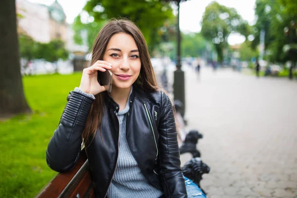 Retrato de una mujer feliz sentada en el banco y hablando por teléfono al aire libre — Foto de Stock