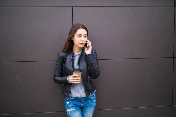 Joven alegre enviando mensajes de texto y sosteniendo café mientras se apoya contra la pared en el exterior urbano — Foto de Stock