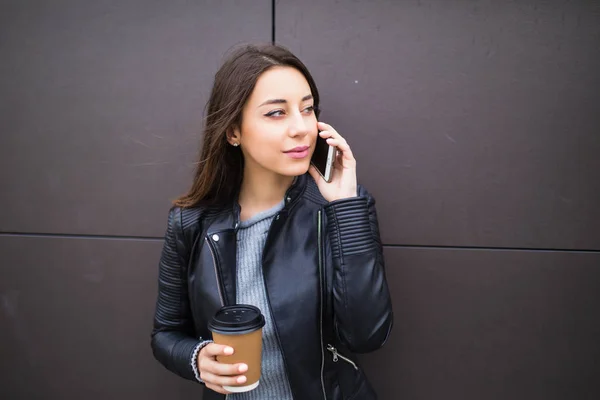 Hermosa estudiante apoyada en la pared, hablando por teléfono inteligente y sosteniendo el café para llevar en papercup, escuchando a su interlocutor con interés, sonriendo — Foto de Stock