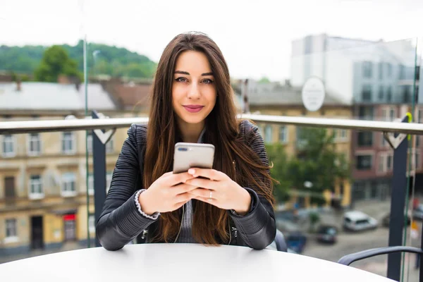 Attractive young brunette woman typing on phone on terrace on big city view background.