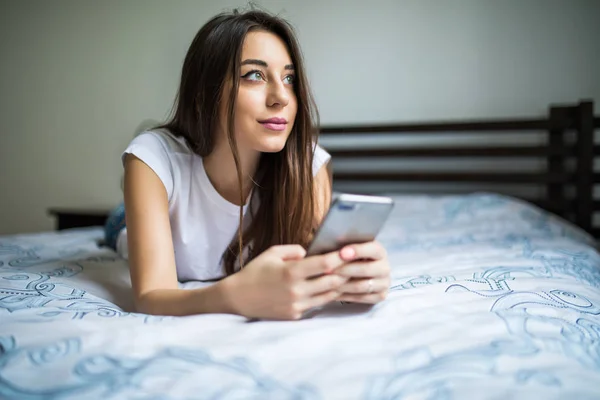 Счастливая женщина, сидящая на кровати и читающая смс на смартфоне. Улыбающаяся девушка, сидящая в спальне в непринужденной беседе с другом в чате. Smiling woman surfing net with cell phone at home . — стоковое фото