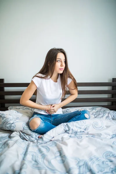 Jovem mulher de beleza que sofre de dor abdominal enquanto se senta na cama em casa — Fotografia de Stock