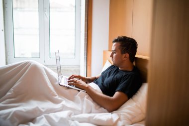 Bir dizüstü bilgisayarda evde çalışma yatakta yatan genç adam