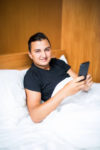 Красивый молодой человек в пижаме, пользующийся сотовым телефоном, лежа утром в постели — стоковое фото