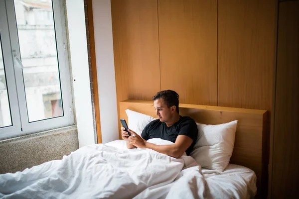 Молодой человек в постели просыпается с сотовым телефоном дома утром — стоковое фото