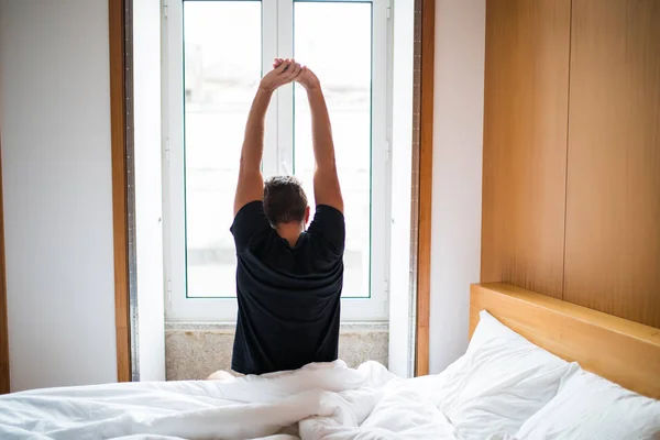 Rear view of a young man waking up in bed and stretching his arms in the morning