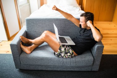 Genç adam--dan laptop mutlu zafer duygu ile iyi bir haber olsun. Yakışıklı genç adam evde kanepede otururken gülümseme ile onun laptop kullanarak yan görünüm