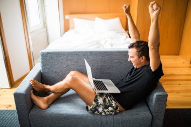 Genç adam--dan laptop mutlu zafer duygu ile iyi bir haber olsun. Yakışıklı genç adam evde kanepede otururken gülümseme ile onun laptop kullanarak yan görünüm