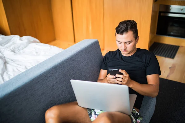 Joven guapo usando su computadora portátil y teléfono mientras está sentado en el sofá en casa — Foto de Stock