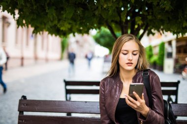 Genç kadın şehirde bankta oturmuş smartphone kullanır.
