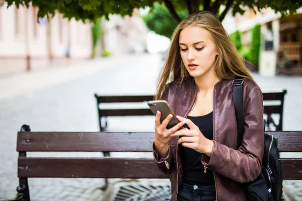 Молодая женщина использует смартфон, сидя на скамейке в городе . — стоковое фото
