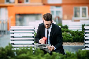 Klasik takım elbiseli yakışıklı sakallı işadamı kahve içme ve şehrin bankta oturan bir gazete okuyor