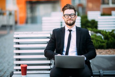 Elinde kahve fincanı ile tahta bankta oturmuş takım elbise ve dizüstü düşünceli genç işadamı kucağına yerleştirilir.