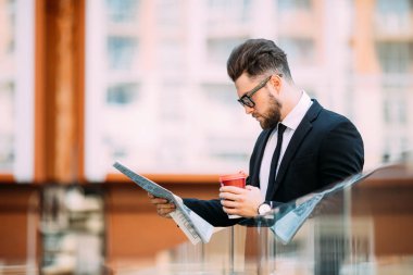 Klasik takım elbiseli yakışıklı sakallı işadamı kahve içme ve ofis Merkezi balkonda istirahat ederken bir gazete tutan