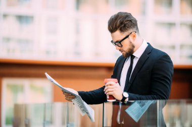 Klasik takım elbiseli yakışıklı sakallı işadamı kahve içme ve ofis Merkezi balkonda istirahat ederken bir gazete tutan