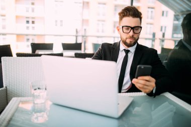 takım elbise dışında bir yerde bir kahve dükkanında cep telefonu yazarak bir teras laptop ile rahatlatıcı yakışıklı iş adamı