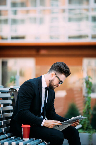 Handsome bearded businessman is reading newspaper, drinking coffee while sitting on bench outdoors