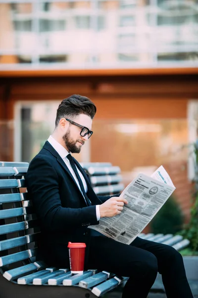 Stilig skäggiga affärsman i klassiska kostym är dricka kaffe och läsa en tidning medan de vilar på bänken i staden — Stockfoto