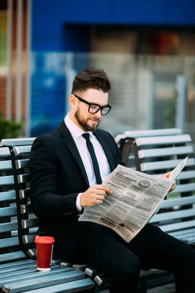 Красивий бородатий бізнесмен читає газету, п'є каву, сидячи на лавці на відкритому повітрі — стокове фото