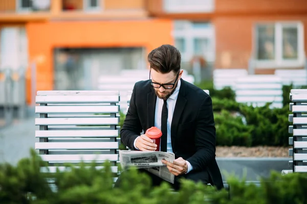 Klasik takım elbiseli yakışıklı sakallı işadamı kahve içme ve şehrin bankta oturan bir gazete okuyor