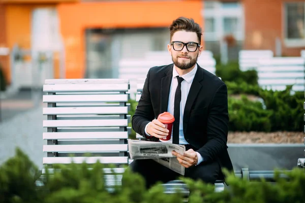 Stilig affärsman läsa tidningen, dricka kaffe, tittar på kameran och ler medan du sitter på bänken utomhus — Stockfoto