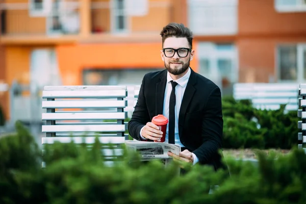 Stilig affärsman läsa tidningen, dricka kaffe, tittar på kameran och ler medan du sitter på bänken utomhus — Stockfoto