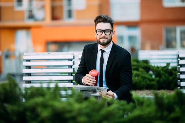 Handsome businessman is reading newspaper, drinking coffee, looking at camera and smiling while sitting on bench outdoors — Stock Photo, Image