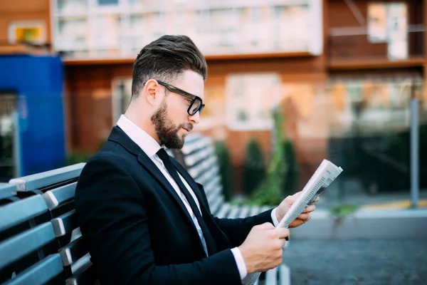 Klasik takım elbiseli yakışıklı sakallı işadamı kahve içme ve şehrin bankta oturan bir gazete okuyor