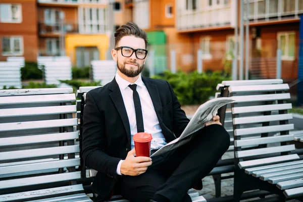 Klasik takım elbiseli yakışıklı sakallı işadamı kahve içme ve şehrin bankta oturan bir gazete okuyor