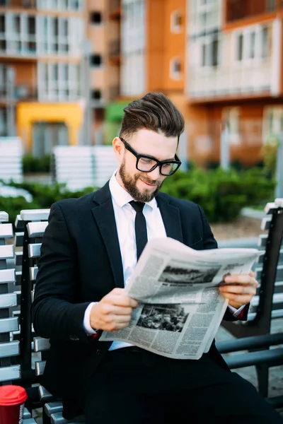 Stilig skäggiga affärsman i klassiska kostym är dricka kaffe och läsa en tidning medan de vilar på bänken i staden — Stockfoto