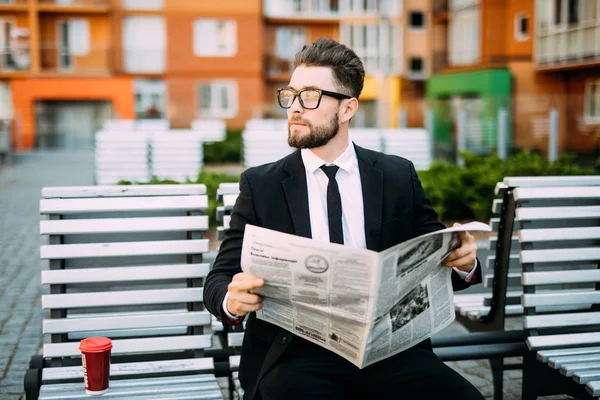 Stilig skäggiga affärsman i klassiska kostym är dricka kaffe och läsa en tidning medan de vilar på bänken i staden — Stockfoto