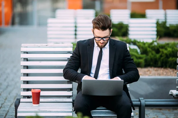 Giovane uomo d'affari premuroso in giacca e cravatta seduto su una panchina di legno con una tazza di caffè in mano e un computer portatile in grembo . — Foto Stock