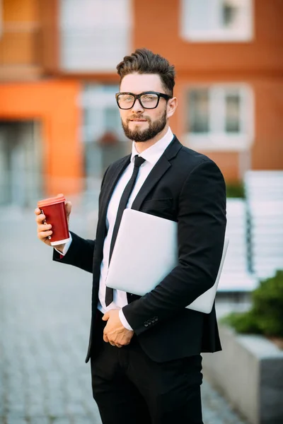 Бізнесмен у костюмі з кавою в руці, що йде з ноутбуком на міській вулиці — стокове фото