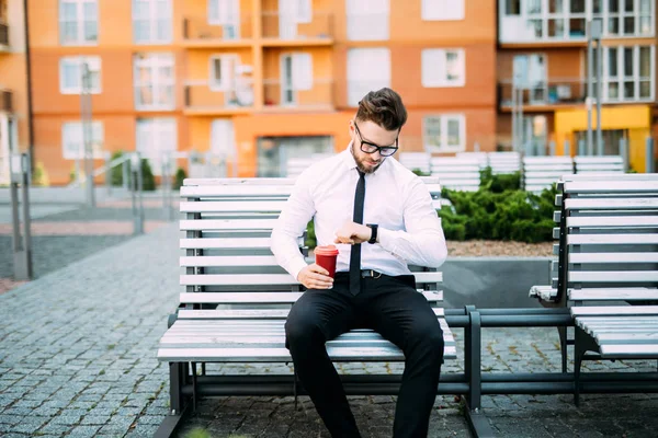 Молодой бизнесмен сидит на скамейке и смотрит на наручные часы, пока пьет кофе. . — стоковое фото