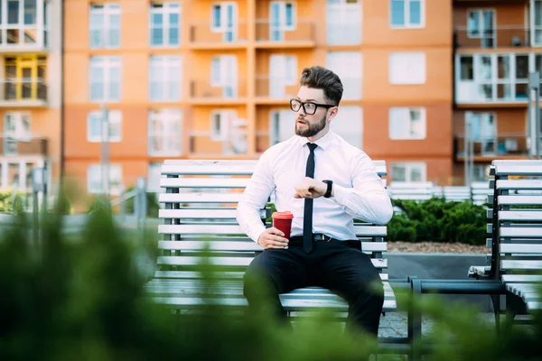 Stilig skäggiga affärsman i klassisk kostym och solglasögon är dricka kaffe och titta på sin klocka medan de vilar på bänken i staden — Stockfoto