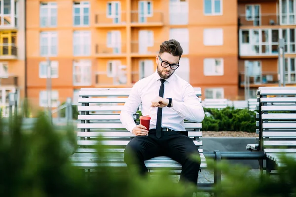 Açık havada bankta oturan ve kol saati drinck kahve süre seyir genç işadamı.