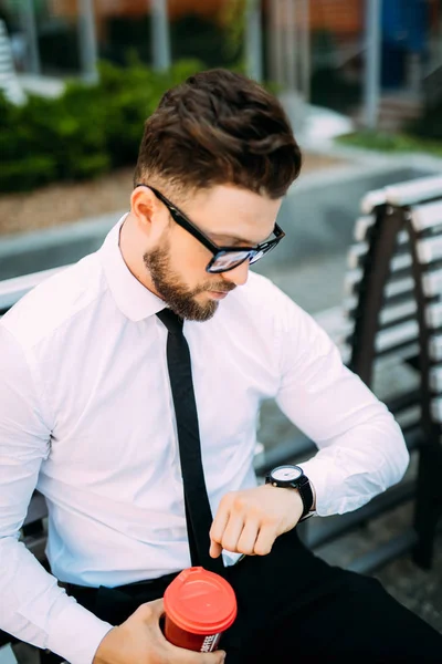 Hombre de negocios barbudo guapo en traje clásico y gafas de sol está bebiendo café y mirando su reloj mientras descansa en el banco de la ciudad — Foto de Stock