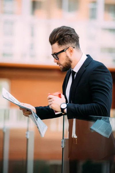 Hezký vousatý podnikatel v klasickém obleku je pití kávy a drží noviny při odpočinku na balkóně office center — Stock fotografie