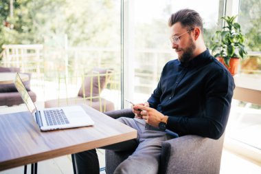 Ofiste cama tabloda, dizüstü bilgisayar ve akıllı telefon kullanan yakışıklı sakallı adam